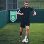 A person wearing a black sports outfit and running shoes is playing soccer on an outdoor field, showcasing the benefits of physical health. They are dribbling a soccer ball and appear focused. The background features a sign with a soccer emblem and the text 