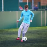 A person wearing a blue long-sleeved shirt and grey sweatpants is playing bubble football on a green, outdoor turf field. They are about to kick a soccer ball, and it appears to be a practice or casual game. The background features a fence and blurred structures. © Powerleague