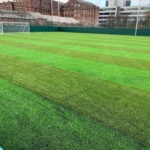 A well-maintained soccer field with bright green artificial turf and clearly marked lines awaits the local football leagues. There are two soccer goals, one at either end of the field. The field is surrounded by a high green fence, and several buildings are visible in the background. © Powerleague