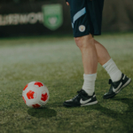 A person in dark athletic shorts and black Nike soccer cleats stands on a grass field next to a red and white soccer ball, seemingly ready for football tournaments. The person's white socks are pulled up to mid-calf. The background is blurred, showing part of a green and black banner. © Powerleague