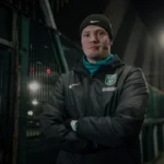 A person stands outdoors at night in front of a fenced area, illuminated by bright lights in the background. They are wearing a black beanie, a black Nike sports jacket with 