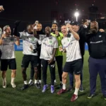 A group of men, all wearing sports attire, stand together on an outdoor soccer field at night. They are smiling, some holding bottled beverages, appearing to celebrate one of their team building days. Bright lights from the cityscape in the background illuminate the scene. © Powerleague