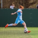 A women's football player in a light blue uniform and red cleats is about to kick a ball on a green field. The player's leg is extended back, and they appear focused on the ball. A dark green fence and spectators in the background are visible. © Powerleague