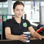 A woman wearing a black polo shirt with a soccer logo stands behind a counter, holding a paper cup. She is smiling slightly, and the background features various items typically found in a café or concession stand, including drink dispensers and plastic cups. Welcome to the home of 5-a-side! © Powerleague