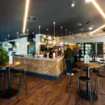 Modern bar interior with an industrial design featuring exposed brick walls and wooden accents. High tables and bar stools are arranged around a central bar counter. Pendant lights hang from the ceiling, and people are seen socializing in the background, much like careers at Powerleague. © Powerleague