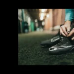 A person is tying the laces of their black Nike athletic shoes, preparing for an intense day of football tournaments. The setting looks like an indoor hallway or corridor with artificial turf flooring and blurred lighting in the background. © Powerleague