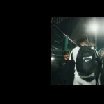 A night-time scene of three young men standing together outdoors near a fence. One person, wearing a black Nike backpack, has their back to the camera. The other two, possibly student athletes, are facing him and smiling in their dark athletic clothing under bright lights from the nearby 5-a-side field. © Powerleague