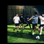 A group of people are playing an intense game of indoor soccer on a green turf field, showcasing their health and stamina. One player in a white shirt and black shorts is attempting to kick the ball while another player in a gray shirt and black shorts is trying to block him. Others are in the background. © Powerleague