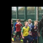 A group of young boys wearing various soccer uniforms enthusiastically follow a smiling man in a black jacket. They are outside near a fenced area, enjoying what appears to be a fun and energetic moment, reminiscent of the lively atmosphere at football holiday camps. © Powerleague