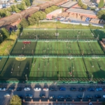 Aerial view of a sports complex featuring several adjacent artificial turf soccer fields buzzing with players from local football leagues. Surrounding the fields are parked cars, adjacent buildings, trees, and a bustling parking lot. The area is bathed in daylight. © Powerleague