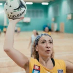 A woman wearing a yellow jersey and a headband is poised to throw a netball in an indoor court, where 5-a-side referees monitor the game. Other players and netball hoops are visible in the background. © Powerleague