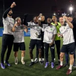 A group of eight men stands on a soccer field at night, celebrating and holding small trophies from football tournaments. They are wearing matching white shirts with 