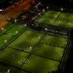 Aerial view of multiple illuminated, fenced soccer fields at night, bustling with players from local football leagues. Adjacent to the fields is a parking lot filled with numerous cars. Surrounding buildings and streetlights are also visible, contributing to the nighttime cityscape. © Powerleague