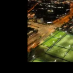 Aerial view of several brightly lit sports fields at night, surrounded by netted fences. Players from local football leagues can be seen engaging in various games on the fields. Nearby, city streets are illuminated by streetlights, with buildings and vehicles visible in the background. © Powerleague