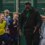 A smiling coach enthusiastically gestures while standing beside a group of children in soccer jerseys. The children are smiling and laughing, displaying team spirit and excitement, as if enjoying one of those vibrant football holiday camps. They are outside, near a green wire fence on a sports field. © Powerleague