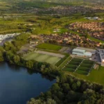 Aerial view of a scenic landscape featuring a tranquil lake surrounded by lush greenery. Nearby, there's a residential area with neatly arranged houses, where local football leagues gather at the sports complex amidst open fields under a clear sky. © Powerleague