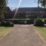 A path leads to a fenced-in sports field under a clear blue sky, where local football leagues compete. Brick buildings and trees stand in the background as sunlight casts a bright beam across the scene. © Powerleague