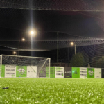 A well-lit soccer field at night with a goalpost in the center hosts local football leagues. The field is surrounded by advertising boards and a net overhead, while stadium lights illuminate the green artificial turf. © Powerleague