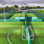 Aerial view of a fenced multi-purpose sports facility featuring artificial turf soccer fields, often bustling with local football leagues. Two small goals are placed on the nearest field. Surrounding trees and cloudy skies add a serene backdrop to the lively scene. © Powerleague