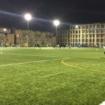 A nighttime soccer match is taking place on a well-lit field. Players from local football leagues are scattered across the field, with some in white jerseys and others in darker jerseys. In the background, there are buildings illuminated by floodlights. A spectator stands near the sideline. © Powerleague