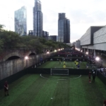 A small soccer field is nestled between tall buildings and a raised structure. Players in yellow and black jerseys, representing local football leagues, move swiftly on the field. Spectators line the perimeter, while lush green trees border one side and skyscrapers rise in the background. © Powerleague