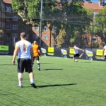 A group of men playing soccer on a sunny day. Representing local football leagues, players in white and orange jerseys compete on a fenced field, surrounded by buildings and trees in the background. © Powerleague