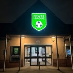 The entrance of the Power League building at night is illuminated by a bright green sign featuring a soccer ball emblem, symbolizing its ties to local football leagues. Through the glass doors, the inviting interior and a welcoming sign on the left wall beckon visitors inside. © Powerleague