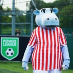 A person in a hippo costume wearing a red and white striped soccer jersey stands on a sports field. In the background, there's a 