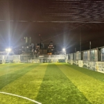 A brightly lit rooftop soccer field at night, featuring artificial grass and surrounded by tall netting, hosts local football leagues. City buildings with illuminated windows are visible in the background. © Powerleague