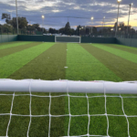 View of a soccer field from behind a goal, under a cloudy sky at dusk. The well-lit turf hosts local football leagues and is surrounded by a green fence. Trees and other urban elements are visible in the background, painting an idyllic scene for community sports enthusiasts. © Powerleague