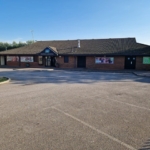 A brick building with a dark roof stands against a clear blue sky, displaying large windows and a 