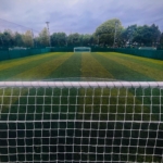 View of a lush green soccer field seen from behind a goal net, often used for local football leagues. The pitch is surrounded by a tall fence and bordered by trees, with another goal visible in the distance under a cloudy sky. © Powerleague