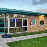 A brick building with a teal roof, featuring a glass entrance and several posters, proudly announces the 