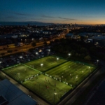 Aerial view of a sports complex at dusk, showcasing two illuminated soccer fields hosting local football league matches. The surrounding cityscape, with buildings and streetlights, comes alive under the darkening sky. © Powerleague