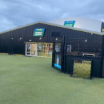 Exterior view of a Powerleague sports facility showcasing a modern building with a glass entrance and black metal facade. The sign above reads 