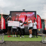 A team of six men, champions of the local football leagues, pose triumphantly with a trophy and a large check on stage. They stand in front of a screen displaying 