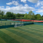 A view of multiple outdoor soccer fields with green artificial turf, ideal for local football leagues. Some fields feature red barriers and small soccer goals. The area is surrounded by trees under a partly cloudy sky, with two people standing near the far fence. © Powerleague