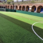 A small outdoor soccer field with artificial turf, ideal for local football leagues, is surrounded by green fencing. Brick arches and a sign with text are visible in the background as sunlight casts shadows across the field. © Powerleague