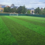 A well-maintained outdoor soccer field with artificial turf, featuring a goal at one end and often hosting local football leagues. The field is surrounded by high netting, with urban buildings visible in the background under a partly cloudy sky. © Powerleague