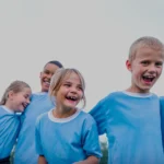 Four children wearing blue shirts are standing close together outdoors, smiling and laughing. The group includes three boys and one girl, all appearing happy and engaged in playful activity at a football holiday camp, with a clear sky in the background. © Powerleague