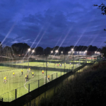 A nighttime scene of an illuminated outdoor sports field hosts local football leagues, where several teams play soccer on multiple pitches. Players in yellow and orange jerseys are visible amid the action. Trees and a dark blue sky serve as the backdrop. © Powerleague