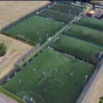 Aerial view of multiple small, fenced soccer fields bustling with local football leagues in action. The surrounding area features lush grass and trees. A building boasting an Adidas logo is visible in the top right corner. © Powerleague