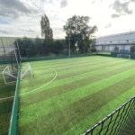 Aerial view of an empty, fenced, artificial grass soccer field under a partly cloudy sky. The field, often bustling with local football leagues, has two goals and is surrounded by trees and a tall building. The alternating light and dark green stripes create a vibrant pattern. © Powerleague