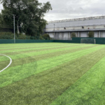 A lush, green soccer field with clear white markings, often hosting local football leagues. There's a single goalpost at one end and, in the background, a tall industrial building and trees beneath an overcast sky. © Powerleague