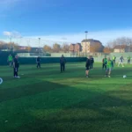 A group of people play soccer on an outdoor artificial turf field under a clear sky, showcasing the spirit of local football leagues. Some players wear green vests, while trees and buildings are visible in the background behind the fenced area. © Powerleague