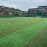 A green artificial soccer field with distinct line markings and a goal at one end hosts local football leagues. The field is surrounded by a tall net fence. In the background, residential buildings stand under a cloudy sky. © Powerleague