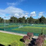 A sports complex with multiple small soccer fields covered in artificial turf, enclosed by green fencing. The fields are outdoors, surrounded by trees under a blue sky with scattered clouds. Catering to local football leagues, the foreground has a well-maintained lawn and decorative shrubs. © Powerleague