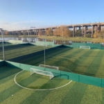 A series of fenced football fields with green artificial turf hosts local football leagues near a river, set against the backdrop of an elevated highway and autumn trees under a clear blue sky. © Powerleague