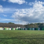 A grassy field hosts a row of green and white banners along a fence, hinting at local football leagues. A building peeks out behind the banners, while trees reach up to meet the blue sky dotted with clouds. © Powerleague