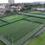 Aerial view of several enclosed soccer fields with green artificial turf buzzes with local football leagues. People are playing on one field, surrounded by residential buildings, trees, and a large stadium in the background. © Powerleague